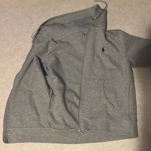 Grå hoodie från Polo Ralph Lauren - Snygg grå hoodie från Polo Ralph Lauren med dragkedja och huva. Klassisk logga broderad på bröstet och två fickor framtill. Perfekt för chill dagar eller när du vill ha en clean streetstyle-look.