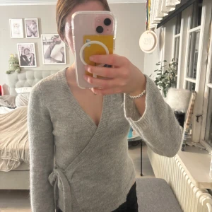 Grå omlottkofta från Gina Tricot - Mysig ljusgrå stickad omlottkofta från Gina Tricot med knyt i sidan och djup v-ringning. Långärmad och ribbade kanter för extra komfort. Perfekt för lager-på-lager under kyliga dagar. Nästan aldrig använd