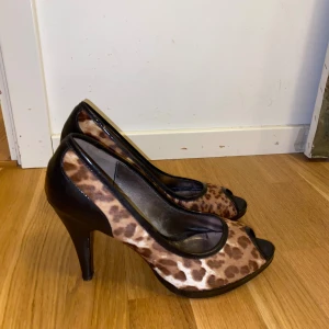 Leopardmönstrade vintage pumps från Tosca Blu - Snygga pumps från Tosca Blu med leopardmönster i bruna och svarta toner. Skorna har öppen tå, hög klack och svarta detaljer i skinn. Perfekta för dig som vill sticka ut med ett djurmönstrat statement.