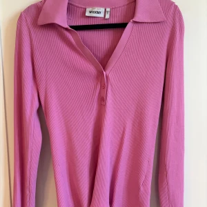 Rosa ribbad långärmad topp Weekday - Säljer en rosa ribbad långärmad topp från Weekday. Toppen har klassisk krage, v-ringning med två knappar framtill och är figurnära i modellen. Perfekt för dig som gillar färg och vill ha en stilren men ändå trendig look. 