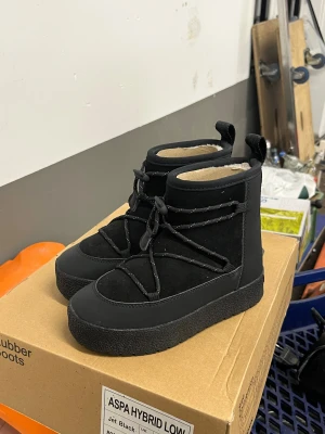 Svarta Aspa Hybrid Low boots - Säljer ett par svarta Aspa Hybrid Low boots i storlek 36. Skorna har en chunky platt sula med grovt mönster, snörning med reflexdetaljer och är tillverkade i mocka och syntet. Insidan är fodrad för extra komfort och värme. Perfekt för dig som gillar stilrena och funktionella boots.