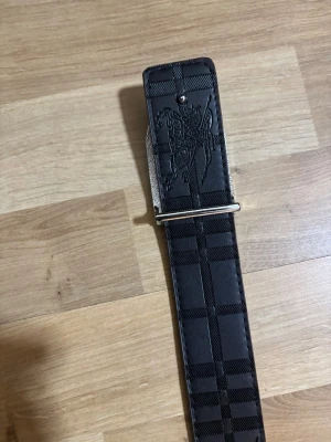 Svart rutig Burberry skärp i skinn - Säljer ett svart Burberry bälte i skinn med klassiskt rutigt mönster och präglad ryttare på spännet. Bältet har en stilren silverfärgad detalj och kommer med original dustbag och box. Perfekt för dig som vill ha en lyxig accessoar med ikonisk design.