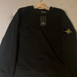 Svart sweatshirt från Stone Island - Svart sweatshirt från Stone Island med klassisk rund hals och ikonisk logotyp-patch på vänster ärm. Tröjan har en clean look och är tillverkad i mjukt bomullsmaterial. Perfekt för dig som gillar streetwear och stilrena plagg.
