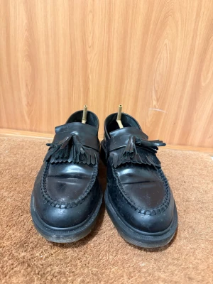 Dr. Martens Tassel Loafers – Svarta – Storlek 43 - Säljer ett par klassiska Dr. Martens Adrian Loafers i svart läder,  storlek 43. Modellen är känd för sin tidlösa design, höga komfort och ikoniska AirWair-sula.  Skick: ✔ Använda men i bra skick ✔ Ytligt slitage och veck på ovansidan (se bilder) ✔ Sulan är använd men fortfarande i gott skick ✔ Inga defekter som påverkar användning  Detaljer: Äkta läder Luftdämpad Dr. Martens-sula Toffelmodell med dekorativa tassels Passar både vardag och mer uppklätt Perfekta om du vill ha ett par stilrena och h