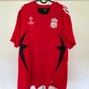 Liverpool Champions League tröja Adidas - Röd Liverpool fotbollströja från Adidas med Champions League-logga och klubbmärke på bröstet. Svarta blixtformade detaljer på sidorna och svart panel på ryggen. Tre vita ränder på axlarna. Tillverkad i polyester, perfekt för dig som älskar fotboll och vill sticka ut.