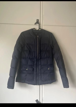 Mörkblå dunjacka med dragkedja - Snygg mörkblå dunjacka med raka linjer och quiltad design. Jackan har rund hals, två fickor med tryckknappar och en smidig dragkedja framtill. Perfekt för kyliga dagar och enkel att matcha med olika outfits.