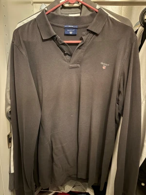 Mörkgrå/brun långärmad pikétröja GANT - Snygg mörkgrå långärmad pikétröja från GANT med klassisk krage och knappar vid halsen. 