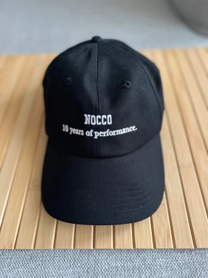  NOCCO svart keps med broderi - Svart keps från NOCCO med vit broderad text 'NOCCO 10 years of performance.' framtill. Klassisk böjd skärm och justerbar rem med metallspänne baktill. Tillverkad i slitstark bomull, perfekt för dig som gillar sportig och clean stil.