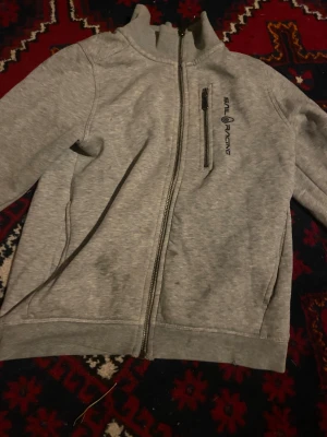 Grå ziptröja från Sail Racing - Grå ziptröja från Sail Racing med hög krage och svart logga på bröstet. Tröjan har hel dragkedja framtill, en bröstficka med dragkedja och två sidofickor. Mjuk och skön, perfekt för lager-på-lager. Klassisk och stilren design.