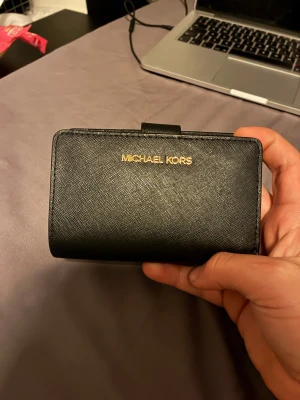 Svart plånbok från Michael Kors - Stilren svart plånbok från Michael Kors i skinn med guldfärgad logga framtill. Plånboken har flera kortfack, myntfack med dragkedja och ett fönster för ID-kort. Perfekt storlek för att ha med sig överallt och snygg detalj i väskan. Köpt för 1000kr aldrig använd