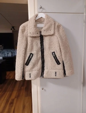 Beige teddyjacka med svarta detaljer - Säljer en beige teddyjacka från Zara med stora krage och svarta dragkedjor på fickor och framtill. Jackan är fluffig och har en loose passform, perfekt för höst och vinter. Materialet är mjuk fleece som ger en mysig vibe. Fickorna med dragkedja är fejkfickor men fickor finns strax ovanför. Mocka inuti. Knappt använd så I mycket bra skick. Normal i storlek.