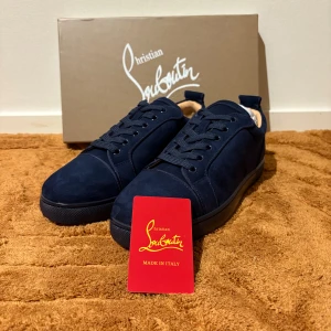 Mörkblå sneakers från Christian Louboutin - Snygga mörkblå sneakers från Christian Louboutin i mjuk mocka med klassisk röd sula och diskret broderad logga på hälen. Skorna har rund tå, platt sula och snörning framtill. Perfekt för dig som gillar exklusiv stil och detaljer.
