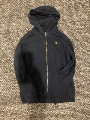 Mörkblå hoodie från Lyle & Scott - Snygg mörkblå hoodie från Lyle & Scott med dragkedja och huva. Tröjan har ett gult broderat örnmärke på bröstet och praktiska fickor framtill. Tillverkad i mjuk bomull, perfekt för chill dagar och sköna outfits.