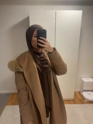Beige Max Mara kappa med päls - Snygg beige kappa från Max Mara Studio med stor fluffig päls på huvan och omlottmodell med bälte i midjan. Kappan har långa ärmar och en klassisk, loose passform som ger en chill och lyxig vibe. Perfekt för dig som gillar stilrena och tidlösa plagg. Den är i storlek L, men passar även folk som har S/M. Den är i ett helt perfekt skick! Köpt för 12 000 kr från Johnells.