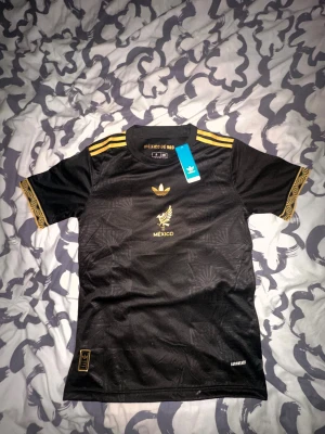 Svart Mexiko fotbollströja Adidas S - Snygg svart Mexiko fotbollströja från Adidas med guldiga detaljer och broderad örn på bröstet. Tröjan har korta ärmar med gulddetaljer och tre guldiga ränder på axlarna. Materialet är lätt polyester med diskret mönster och Aeroready-teknologi för extra komfort.