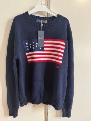 Mörkblå stickad tröja Polo Ralph Lauren - Mörkblå stickad tröja från Polo Ralph Lauren med amerikansk flagga i rött, vitt och blått på bröstet samt RL-broderi. Klassisk rund halsringning och ribbade muddar. Perfekt för dig som gillar preppy och tidlös stil.