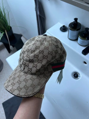 Gucci beige keps med GG-mönster - Snygg beige keps från Gucci med klassiskt GG-mönster över hela kepsen och grönt/rött band på sidorna. Justerbar rem baktill i skinn och insida i brunt tyg. Perfekt för dig som vill ha en lyxig och trendig accessoar. Får påse till
