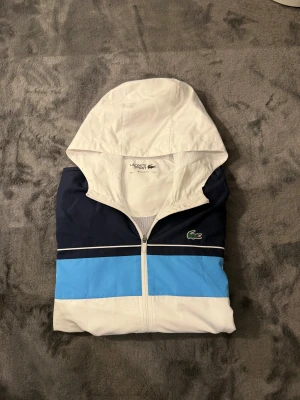 Lacoste Sport blå-vit vindjacka - Snygg vindjacka från Lacoste Sport med huva, dragkedja och elastiska muddar. Jackan har färgblock i vitt, marinblått och ljusblått samt den klassiska Lacoste-krokodilen på bröstet. Perfekt för dig som gillar sportig och clean stil. Obs har två små små hål vid ryggen man ser kanapt vid användning 