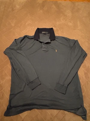 Mörkblå/grå långärmad pikétröja Polo Ralph Lauren - Bra julklapp!!! Snygg mörkblå långärmad pikétröja från Polo Ralph Lauren med klassisk krage i svart och den ikoniska broderade loggan på bröstet. Tröjan är tillverkad i mjuk bomull och har muddar vid ärmsluten för en schysst passform.  (🔥om det syns några fleckar så är det sånt som går att tvätta 🧽 🔥)