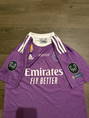 Real Madrid Ronaldo 7 lila fotbollströja - Säljer en lila Real Madrid fotbollströja från Adidas med Ronaldo 7 på ryggen. Tröjan har vita detaljer, korta ärmar och tryck från Champions League-finalen 2017 i Cardiff. Materialet är lätt polyester med klassiska Adidas-ränder på axlarna och klubbmärke på bröstet.