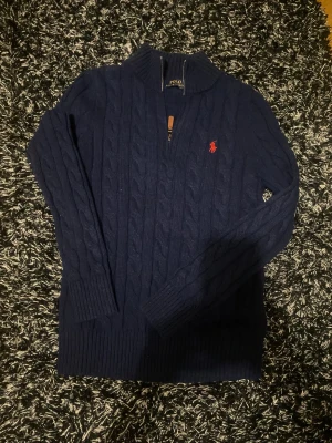 Marinblå stickad tröja Polo Ralph Lauren - Snygg marinblå stickad tröja från Polo Ralph Lauren med kabelstickat mönster och halv dragkedja vid halsen. Tröjan har långärmad passform, ribbade muddar och klassisk röd logga broderad på bröstet. Perfekt för dig som gillar stilren och tidlös design.