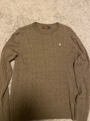 Brun kabelstickad tröja Morris - Snygg brun kabelstickad tröja från Morris i 100% merinoull. Rund hals och diskret broderad logga på bröstet. Perfekt för dig som gillar klassisk stil med en modern touch. Långärmad och mjuk, passar dig som vill ha något bekvämt och stilrent. Passar storlek 164-170