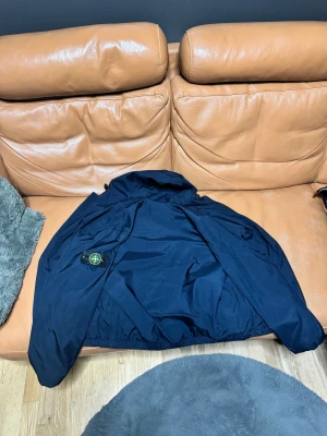 Mörkblå vindjacka från Stone Island - Snygg mörkblå vindjacka från Stone Island med klassisk patch på ärmen. Jackan har huva, dragkedja framtill och sidofickor med zip. Materialet är lätt och syntetiskt, perfekt för blåsiga dagar. Stilsäker och enkel att matcha med jeans eller cargo.