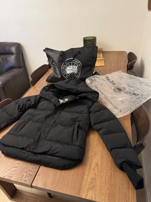 Canada goose macmillan jacka - Säljer en svart pufferjacka från Canada Goose med ikonisk patch på ärmen och huva. Jackan har dragkedja framtill och stora fickor. Tillverkad i slitstarkt material och fodrad med dun för extra värme. Perfekt för kalla dagar och riktigt snygg streetstil. Finns äkthets bevis