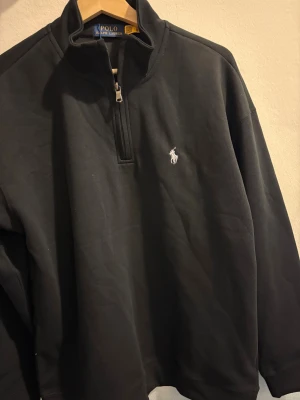 Svart half zip tröja Polo Ralph Lauren - Svart långärmad tröja från Polo Ralph Lauren med half zip och klassisk broderad logga på bröstet. Tröjan har en hög krage och är gjord i mjuk bomull, perfekt för chill dagar. Snygg och enkel att matcha med jeans eller joggers.