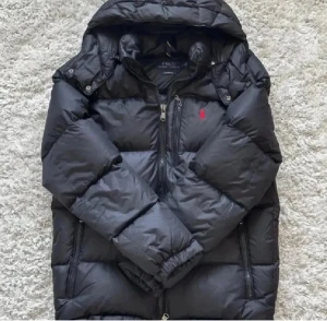 Svart pufferjacka Polo Ralph Lauren - Svart pufferjacka från Polo Ralph Lauren med huva, flera dragkedjor och ikonisk röd logga på bröstet. Jackan har en quiltad design, elastiska muddar och praktiska fickor med dragkedja. Perfekt för kalla dagar och har en clean, modern look.