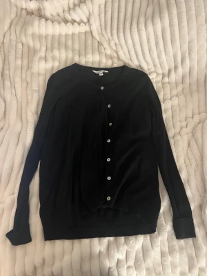 Svart kofta från BASIC U - En stilren svart kofta från BASIC U med rund halsringning och knäppning framtill med vita knappar. Långärmad modell i mjukt material, perfekt att slänga över en t-shirt eller linne. Enkel och klassisk design som funkar till det mesta.