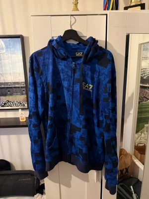 Blå camomönstrad hoodie från EA7 - Säljer en blå hoodie från EA7 Emporio Armani med svart camomönster och gul logga på bröstet. Tröjan har dragkedja, huva med snörning och två fickor framtill. Perfekt för dig som gillar streetwear och sportig stil.