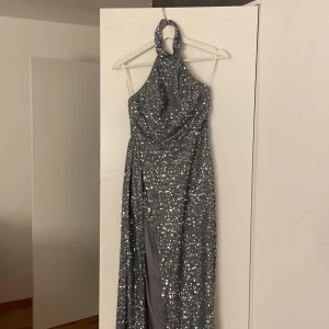 Glittrig silver långklänning med slits - Säljer en maxad långklänning i silver med glittriga paljetter över hela tyget. Klänningen har halterneck, hög slits framtill och en snygg, figurnära passform. Perfekt för dig som vill sticka ut på bal eller nyår. Materialet är mjukt och följsamt.