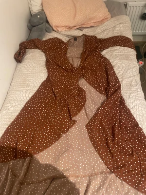 Brun prickig långklänning från SHEIN - Säljer en brun långklänning från SHEIN med vita prickar. Klänningen har långa ärmar, omlottmodell och volangdetaljer längs nederkanten. Tunt och lätt material som ger ett flowy intryck. Perfekt för dig som gillar retrovibbar och mönster!