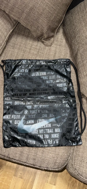 Svart Nike gympåse med tryck - Säljer en svart gympåse från Nike med snörstängning och coolt tryck med text på flera språk. Stor Nike-logga framtill. Perfekt för att bära träningskläder eller skor. Tillverkad i lätt syntetmaterial och har klassisk sportig look.