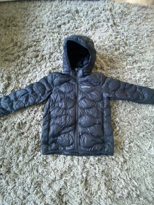 Svart dunjacka Peak Performance 160 - Snygg svart dunjacka från Peak Performance i storlek 160. Jackan har huva, dragkedja framtill och ett quiltat mönster som ger en cool look. Perfekt för kalla dagar och har en stilren design med logga på bröstet.