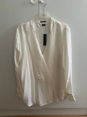Vit blus från Theory - Snygg vit omlottblus från Theory i mjukt och lyxigt silkesmaterial. Blusen har lång ärm, v-ringad hals och knappar vid ärmslut. Stilren och elegant design som ger en clean look. Perfekt för dig som gillar minimalistisk stil och vill ha något som känns exklusivt
