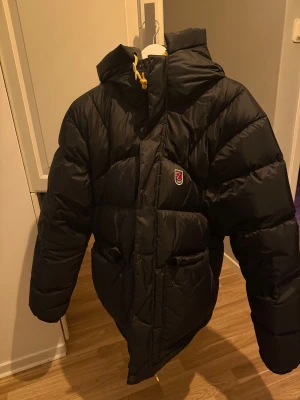 Svart Fjällräven Expedition dunjacka M - Svart Fjällräven dunjacka med huva och gula snören. Jackan har broderad logga på bröstet, stora fickor framtill och dragkedja med tryckknappar. Perfekt för kalla dagar och riktigt snygg streetstil.