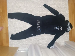 Svarta Hoodrich mjukisbyxor och tröjan - Svarta mjukisbyxor och tröjan från Hoodrich med stor vit broderad logga på framsidan och blå detaljer. Byxorna och tröjan har en loose passform och är tillverkade i mjukt bomullsmaterial. Perfekt för chill eller streetwear-stil. Bokstaven H på byxorna i framsidan är lite suddad annars är den perfekt. Ni kan skriva pågående tracksuiten om ni vill veta mer. Inga defekter har inte märkt eller sett. Använt lite grann annars är den bra, priset kan diskuteras!.