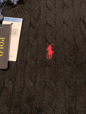 Svart kabelstickad tröja Polo Ralph Lauren -  Pris kan diskuteras.Svart kabelstickad tröja från Polo Ralph Lauren i storlek M. Klassisk rund hals och långärmad modell med röd broderad logga på bröstet. Tröjan är gjord i mjuk ull och har ribbade muddar vid ärmslut och nederkant. Perfekt för dig som gillar stilren och tidlös design.