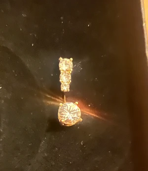 Guldpläterad rostfri navelpiercing med kristaller - Snygg navelpiercing i guldpläterad metall med två glittrande klara kristaller. Den övre delen har två mindre stenar och den undre en större rund kristall som fångar ljuset. Perfekt för dig som vill ha lite extra bling på magen. 