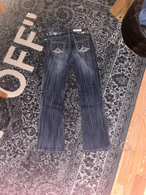 Blå jeans med broderi och dragkedjor - Snygga blå jeans med vita broderier på både fram- och bakfickor samt coola dragkedjor och stjärndetaljer på bakfickorna. Jeansen har en klassisk passform med raka/bootcut ben och är tillverkade i denim. Perfekta för dig som gillar detaljer och unik stil.