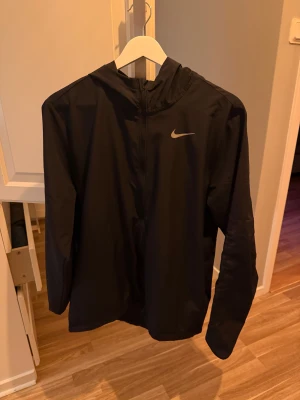 Svart Nike vindjacka Dri-Fit M - Svart Nike vindjacka med huva och dragkedja, perfekt för löpning. Dri-Fit material som andas och håller dig torr. Snygg minimalistisk design med Nike-logga på bröstet. Långärmad och lätt, passar dig som gillar sportig stil.