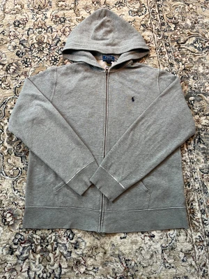 Grå hoodie från Polo Ralph Lauren XL - Säljer en stilren grå hoodie från Polo Ralph Lauren i storlek XL. Tröjan har dragkedja, huva och två fickor framtill. Klassisk broderad logga på bröstet och mjukt bomullsmaterial som är skönt mot huden.Vid slutet av ärmarna är den lite sliten men annars är det inget fel på tröjan. Passar folk runt 165 till 175cm. 