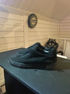 Svarta sneakers från Axel Arigato - Svarta sneakers från Axel Arigato med stilren design och platt sula. Skorna är tillverkade i mocka och har en rund tå. De har en diskret logga på plösen och är helt svarta, vilket ger en clean och modern look. Perfekta för dig som gillar minimalistisk stil. Dom är i bra skick behöver endast tvättas väldigt lite.