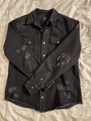 Svart overshirt från Zara med slitningar - Svart overshirt från Zara med coola slitningar och rå kant nedtill. Skjortan har två bröstfickor med knappar, klassisk krage och tryckknappar framtill. Materialet är kraftig bomull som ger en schysst streetkänsla. Perfekt för dig som gillar en edgy stil.