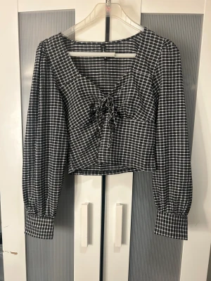 Svartvit rutig blus med snörning - Snygg svartvit rutig blus från H&M med långärmade puffärmar och breda manschetter. Toppen har en croppad passform och v-ringning med snörning framtill. Perfekt för dig som gillar trendiga detaljer och klassiskt rutmönster.