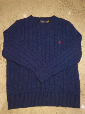 Marinblå kabelstickad tröja Polo Ralph Lauren - Kabelstickad marinblå tröja från Polo Ralph Lauren med klassisk rund hals och röd broderad logga på bröstet. Tröjan har långärmad passform och ribbade muddar vid ärmslut och nederkant. Perfekt för dig som gillar stilren och tidlös design.