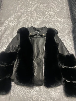 Svart skinnjacka med pälsdetaljer - Snygg svart skinnjacka från Fashion i storlek M med mjuka pälsdetaljer längs ärmar, fram och insida. Jackan har sned dragkedja och krage med tryckknappar. Perfekt för dig som vill ha en unik och varm look med lyxig känsla.