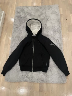 Svart teddyjacka från Moose Knuckles - Svart teddyjacka från Moose Knuckles med vit fluffig fuskpäls på insidan av huvan och framstycket. Jackan har dragkedja, snörning i huvan, två stora fickor fram och en snygg logotyp på ärmen. Perfekt för kalla dagar och har en cool, urban vibe.